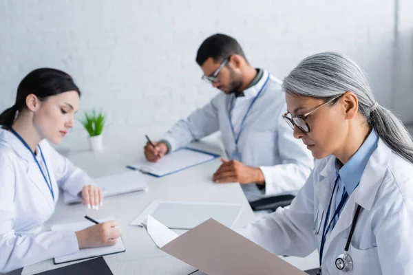 Maduro asiático médico buscando en papel carpeta cerca borrosa colegas trabajando con papeles - foto de stock