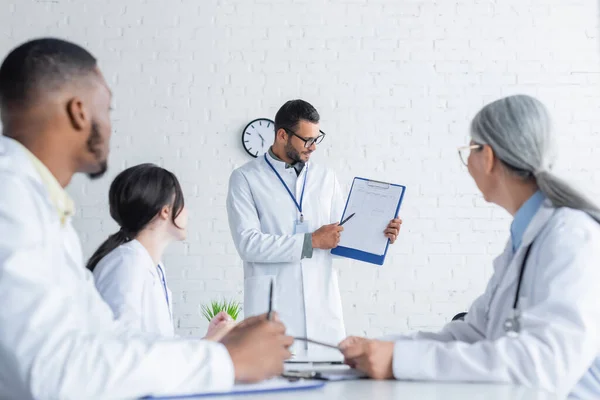 Joven médico apuntando a la tarjeta médica en el portapapeles cerca borrosa colegas multiculturales - foto de stock