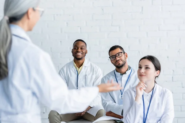 Médico de mediana edad borrosa señalando con la mano cerca de los médicos jóvenes y positivos - foto de stock