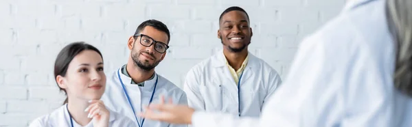 Médicos multiétnicos sorrindo perto de colega maduro falando em primeiro plano borrado, banner — Fotografia de Stock