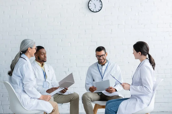 Joven médico con papeles sonriendo cerca de colegas multiétnicos durante el consejo médico - foto de stock