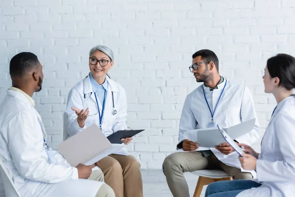 Smiling asian doctor pointing at african american colleagues during medical council — Stock Photo