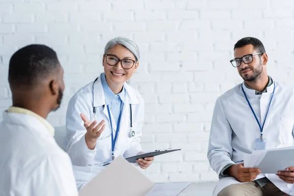 Smiling asian doctor pointing at blurred african colleague during discussion — Stock Photo