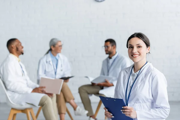 Young doctor with clipboard smiling at camera while multiethnic colleagues talking on blurred background — Stock Photo