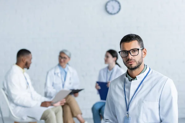 Young physician in eyeglasses looking at camera while multiethnic doctors talking on blurred background — Stock Photo