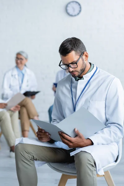 Young doctor in eyeglasses looking in paper folder near blurred colleagues — Stock Photo