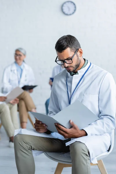 Joven médico en gafas que buscan en la carpeta de papel cerca de colegas borrosos - foto de stock