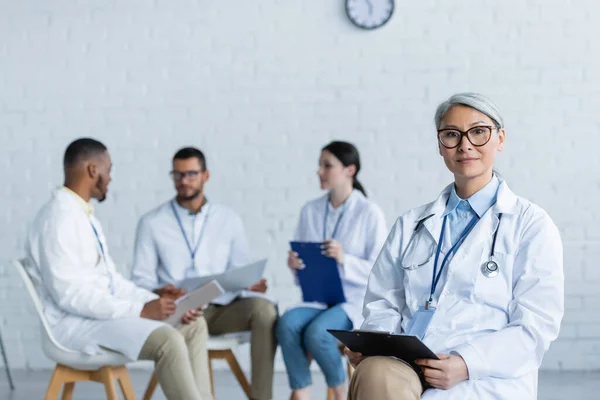 Médico asiático de mediana edad con portapapeles mirando la cámara cerca de colegas multiétnicos hablando sobre fondo borroso - foto de stock