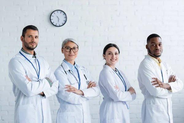 Щасливі мультикультурні лікарі в білих пальто, що стоять з схрещеними руками і дивляться на камеру — стокове фото