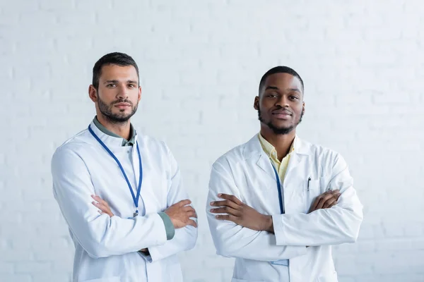 Jóvenes médicos interracial en batas blancas de pie con los brazos cruzados y mirando a la cámara - foto de stock