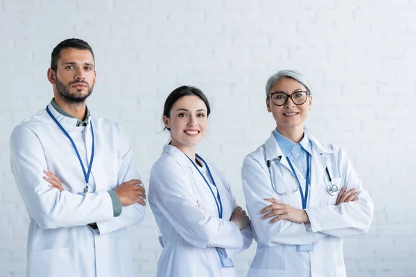 Glückliche multiethnische Ärzte in weißen Mänteln, die mit verschränkten Armen in die Kamera schauen — Stockfoto