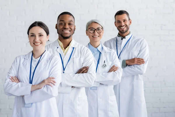 Médicos multiétnicos alegres de pé com braços cruzados e sorrindo para a câmera — Fotografia de Stock