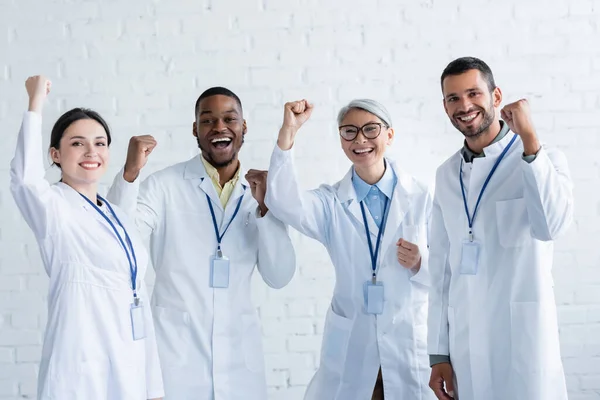 Médicos multiculturales excitados en batas blancas con etiquetas con el nombre mostrando gesto de regocijo - foto de stock