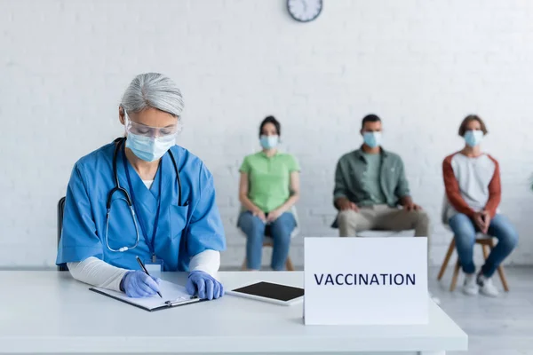Infirmière mature dans l'écriture de masque médical sur presse-papiers près plaque de signe de vaccination et les patients flous — Photo de stock