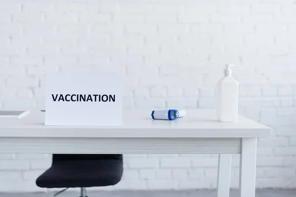 Dispenser bottle with hand sanitizer near pyrometer and sign plate with vaccination lettering on desk — Stock Photo