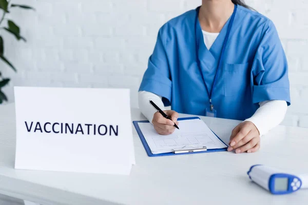 Visão cortada do imunologista escrevendo no cartão médico perto de letras de vacinação e pirômetro borrado — Fotografia de Stock