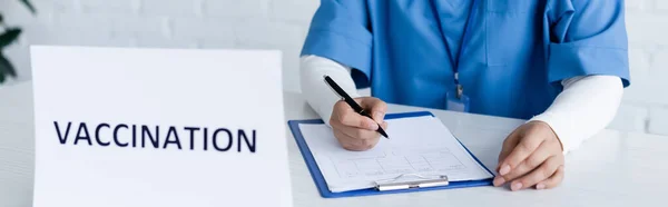 Vista parcial de la escritura del inmunólogo en la tarjeta médica cerca de la inscripción de la vacunación en el escritorio, bandera - foto de stock