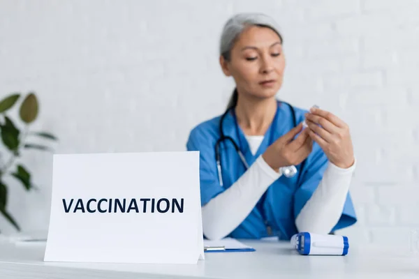 Mature asian doctor holding jar with vaccine near card with vaccination lettering — Stock Photo