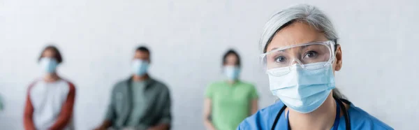 Asiático médico en máscara médica y gafas mirando cámara cerca borrosa pacientes, concepto de vacunación, bandera - foto de stock