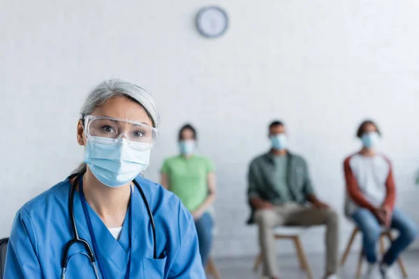 Mature asian nurse in medical mask and goggles looking at camera near blurred patients in vaccination center — Stock Photo