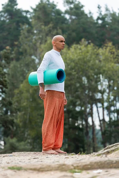 Vista a tutta lunghezza del buddista in felpa e pantaloni harem in piedi con tappetino yoga nella foresta — Foto stock