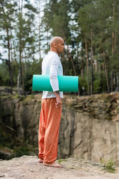 Vista completa de budista de pie con esterilla de yoga en acantilado rocoso en el bosque - foto de stock