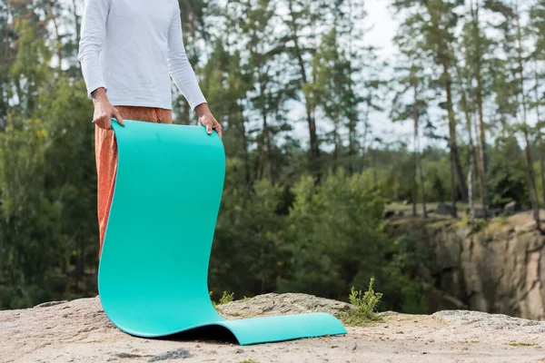 Partial view of buddhist holding yoga mat on rocky cliff in forest — Stock Photo