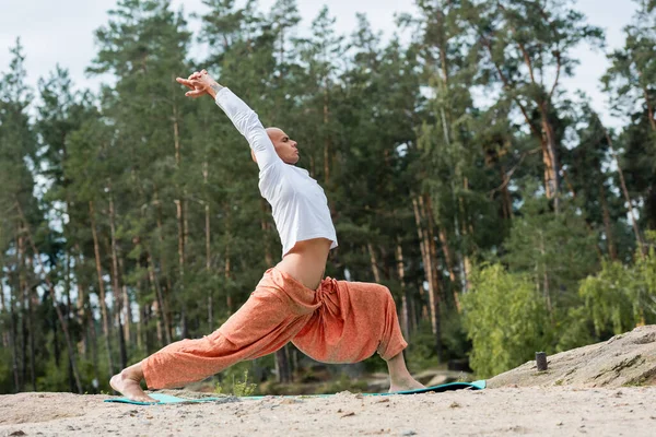 Vista lateral de budista practicando yoga en pose guerrera con las manos levantadas en el bosque - foto de stock