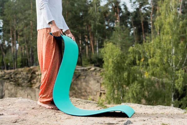 Cropped view of barefoot buddhist holding yoga mat on rocky cliff in forest — Stock Photo