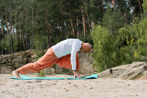 Visão lateral de budista em calças harém praticando crescente pose lunge no tapete de ioga na floresta — Fotografia de Stock