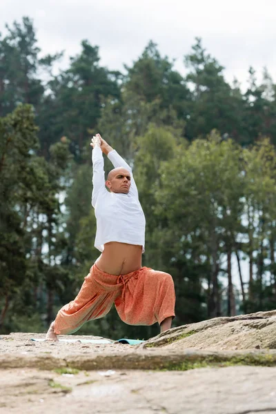 Buddhist en pantalones de harén meditando en pose guerrera con las manos levantadas al aire libre - foto de stock