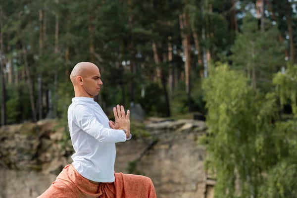 Vista lateral del budista en sudadera blanca meditando con las manos rezando en el bosque - foto de stock