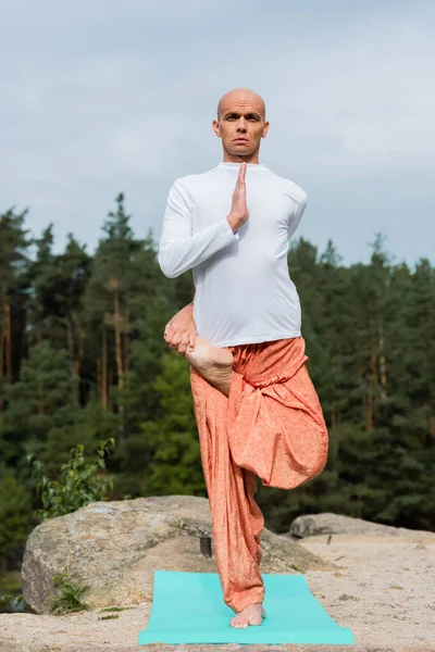 Vista completa de budista en sudadera y pantalones de harén practicando yoga en una pose de árbol con piernas - foto de stock