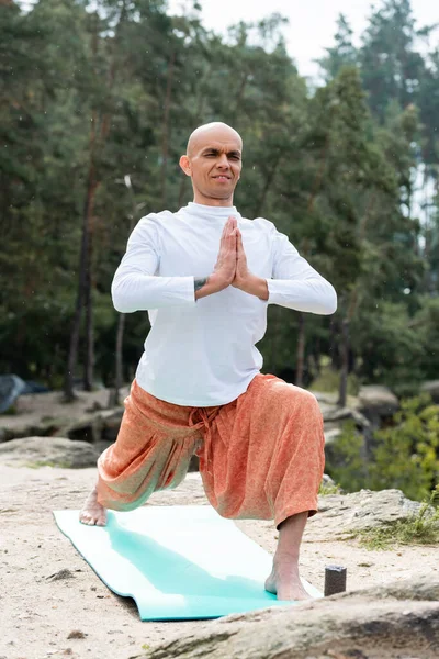 Buddhist en sudadera blanca practicando yoga en pose guerrera con las manos orantes en el bosque - foto de stock