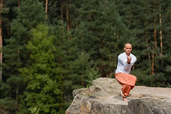 Buddhist in white sweatshirt and harem pants meditating on rocky cliff in yoga pose — Stock Photo