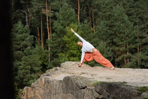 Buddhist in sweatshirt and harem pants practicing yoga in side lunge pose on rocky cliff — Stock Photo