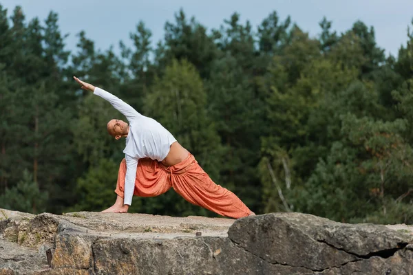 Buddhist en sudadera blanca y pantalones de harén practicando lado embestida pose al aire libre - foto de stock