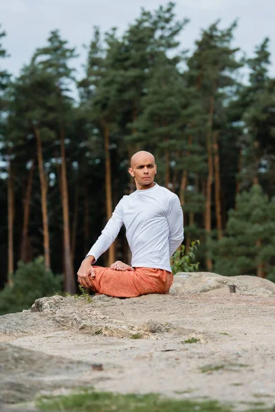 Buddhist en sudadera blanca mirando hacia otro lado mientras practica la pose de loto en un acantilado rocoso - foto de stock