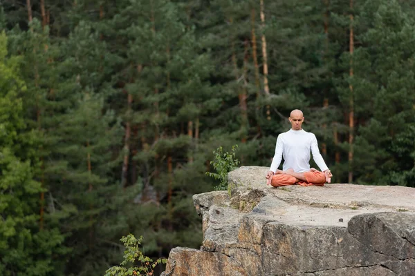 Buddhist in white sweatshirt meditating in lotus pose on rock in forest — Stock Photo