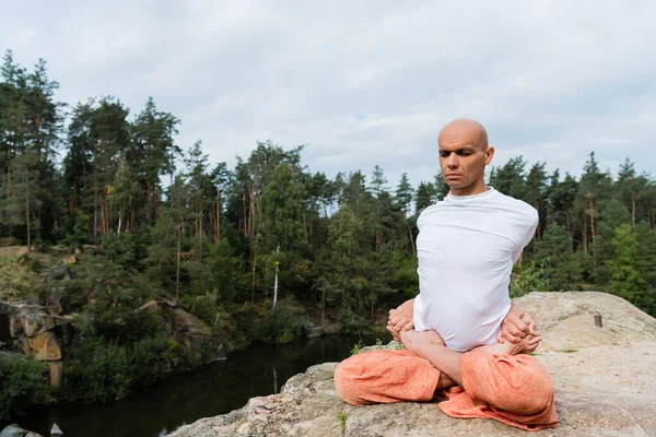 Buddhist en sudadera blanca practicando pose de loto extendido sobre roca en bosque - foto de stock
