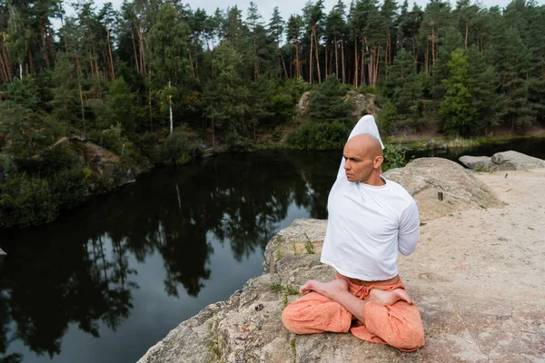 Budista em camisola branca meditando em pose de lótus com as mãos atrás das costas em penhasco rochoso sobre o rio — Fotografia de Stock