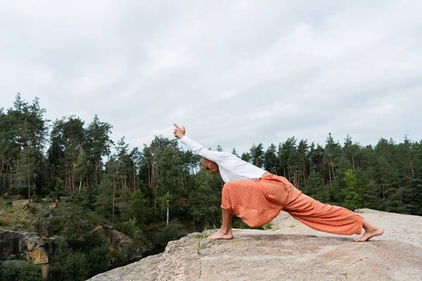 Vista lateral de buddhist meditando en crescent embestida pose en roca en bosque - foto de stock