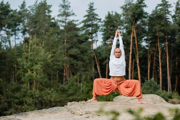 Buddhist en sudadera blanca practicando pose de diosa con las manos levantadas sobre un acantilado rocoso en el bosque - foto de stock
