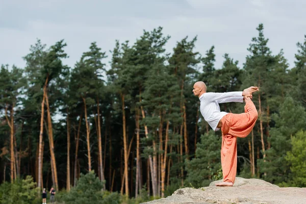 Side view of buddhist practicing standing bow pose on rocky cliff in forest — Stock Photo