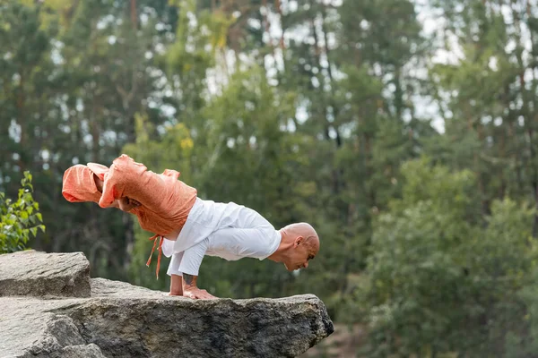 Visão lateral de budista em calças harém meditando em pose de lótus levantada em penhasco rochoso — Fotografia de Stock