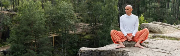 Buddhist en sudadera blanca y pantalones harem meditando sobre roca en bosque, bandera - foto de stock