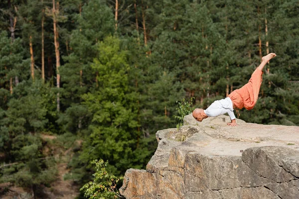 Buddhist in Haremshose übt Armbalancieren auf felsiger Klippe im Wald — Stockfoto