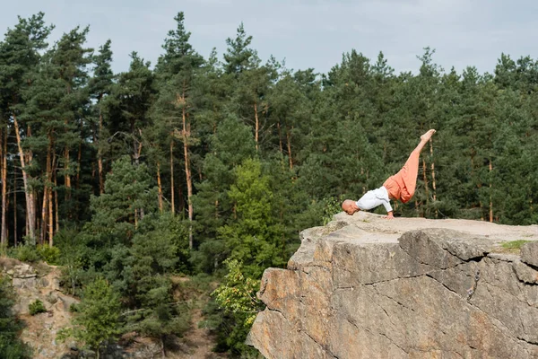 Vista lateral de la postura de equilibrio del brazo practicante budista sobre un alto acantilado rocoso en el bosque - foto de stock