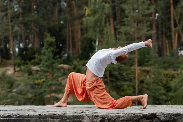 Vista lateral de budista en sudadera y pantalones de harén practicando crescent lunge pose al aire libre - foto de stock