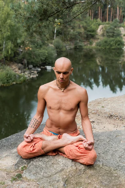 Shirtless, tattooed buddhist meditating in lotus pose on rocky cliff over lake — Stock Photo